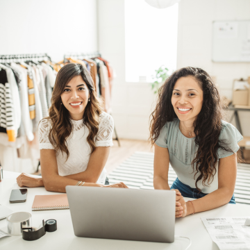 Two female fashion business owners smiling and sitting at a desk