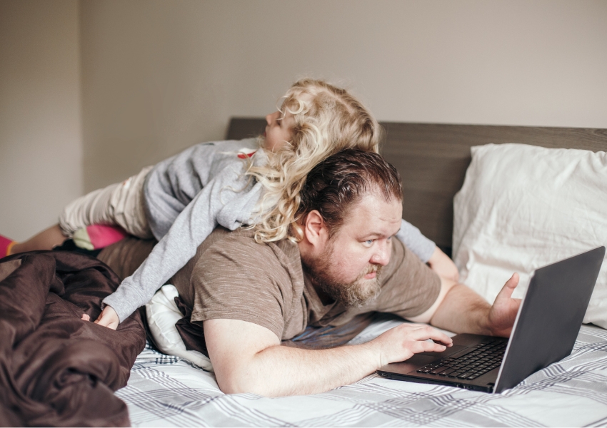 Father on bed looking at laptop with daughter lying on his back