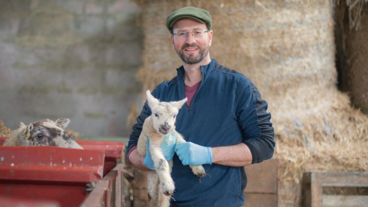 A farmer holds a lamb in one of his farm buildings